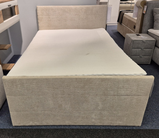 Das beigefarbene Boxspringbett „Elba“ (140 x 200) mit weißer Matratze, daneben ein grauer Nachttisch und ein Sessel.