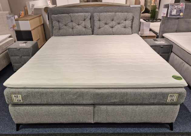 Das Boxspringbett „Cher“ in Grau mit weißer Matratzenauflage und zwei Nachttischen, ausgestellt in einem modernen Ausstellungsraum.