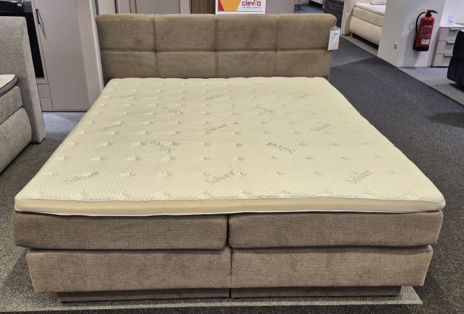 Boxspringbett „Paris“ in Taupe mit wendbarer Taschenfederkernmatratze, grauer Teppich, Möbel im Hintergrund.