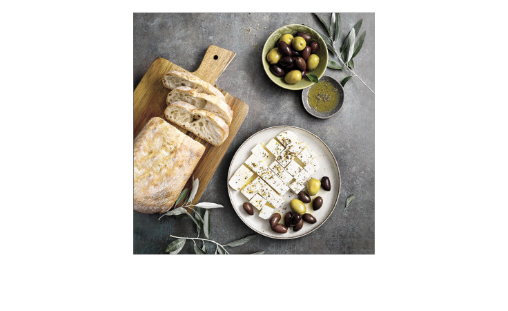 Das Pro-Art Glasbild Mediterrean Food 20x20 cm zeigt Brot, Kräuterfeta im Glas und bunte Oliven auf einem grauen Tisch.