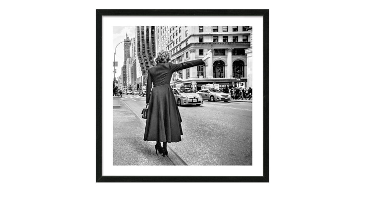 Auf dem Pro-Art Rahmenbild Call A Taxi 52,5 x 52,5 cm ruft eine Frau in einem langen Mantel ein Taxi inmitten hoher Stadtgebäude an.