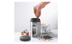 Eine Hand drückt den GEFU Kaffeepad-Dose Piero-Kolben auf eine Kaffeedose neben Pads, Bohnen und einer Kaffeetasse.