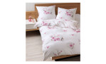 Ein Bett mit Seersucker-Bettwäsche Tango und rosa Blumenkissen neben einem hölzernen Nachttisch mit einer Vase mit Blumen.