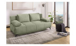 Das Schlafsofa „Giulia“ in Hellgrün mit Schlaffunktion auf einem grauen Teppich, Pflanzen, einer Lampe und großen Fenstern in einem hellen Raum.