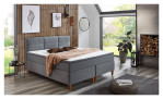 Boxspringbett Kansas in Grau mit einem Mikrofaserbezug und Füße in Eichen-Look. Auf einem dekorierten Hintergrund.