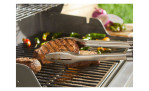 Weber Precision Grillzange aus Edelstahl mit Soft-Touch-Griff hält ein Steak über dem Grill mit Gemüse im Hintergrund.